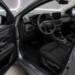 Innenraum eines Dacia Jogger A/T 7-Sitzers von 2024 mit geöffnetem Fahrertür, Lenkrad, Armaturenbrett und Automatik-Wählhebel